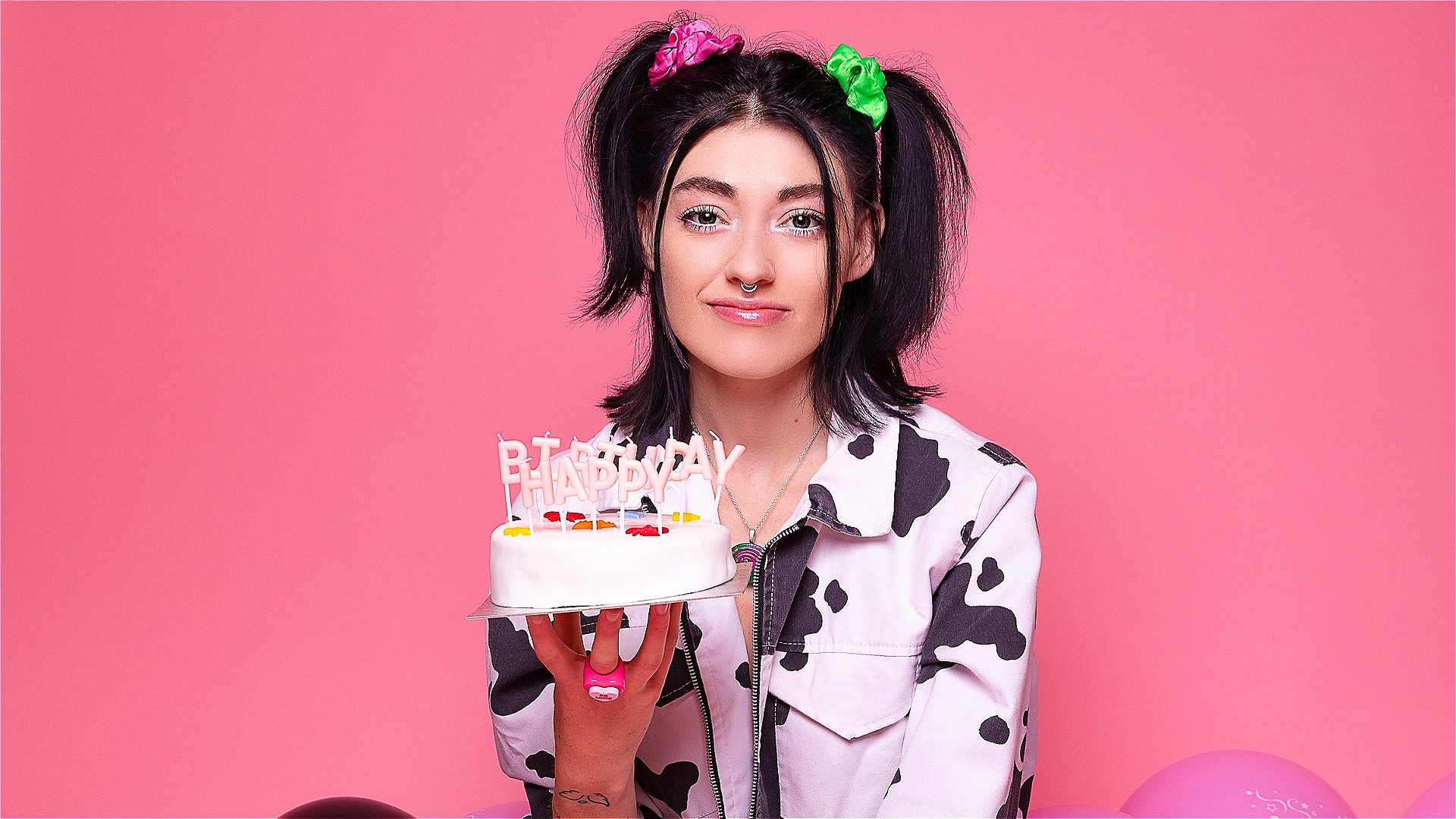 A girl sat on the floor, surrounded by balloons, holding up a birthday cake, with a sarcastic smile on her face.