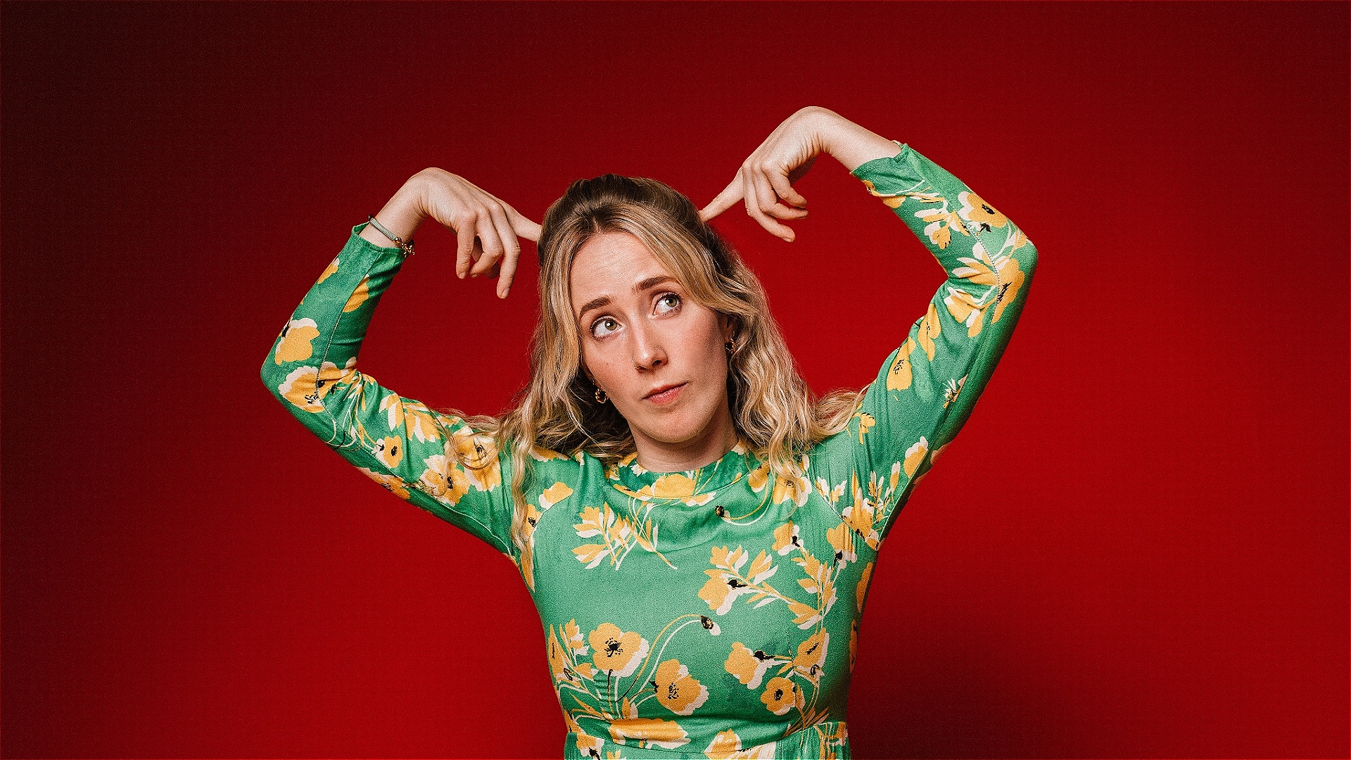 A person in a green floral top, posing with hands above their head in front of a red background.